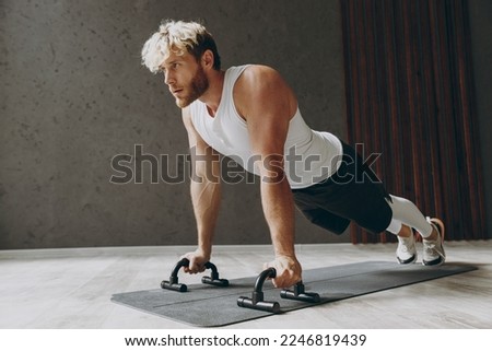 Full body side profile view young strong sporty athletic sportsman man wear white tank shirt doing plank exercise push-ups using handles on floor warm up training indoor at gym. Workout sport concept Royalty-Free Stock Photo #2246819439