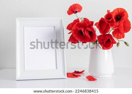 White photo frame on table and red poppies flowers in vase on white wall background.Front view. Place for text, copy space, mockup