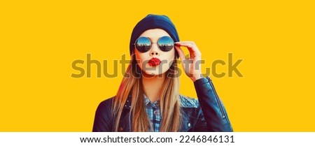 Portrait of stylish beautiful blonde woman blowing her red lips sending sweet air kiss wearing black rock style on yellow background