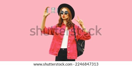 Portrait of modern stylish young woman taking selfie with smartphone blowing her lips sends kiss wearing jacket, black round hat on pink background