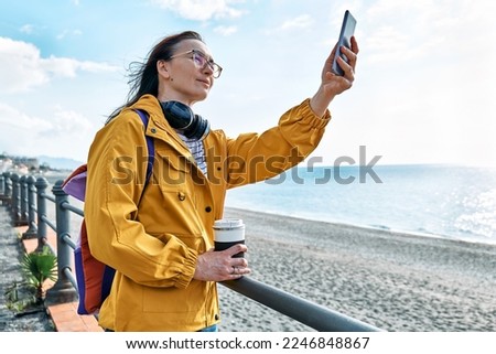 Woman with backpack taking photo of the sea on smartphone on seaside promenade. Relaxing outdoors. The concept of hiking and travel in the low season.