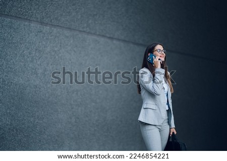 Happy successful business woman using smartphone while walking outside. Beautiful young woman in office suit talking on a mobile and holding a business bag. Legal expert concept. Gray background.