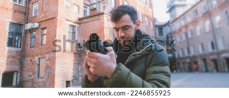 Portrait of a young male photographer with a camera takes a photo or video on the street, in an old courtyard. A handsome guy with a professional camera on the background of an old brick building