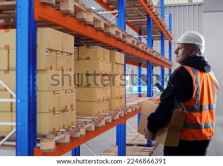 Man is storekeeper. Warehouse worker with box and smartphone. Guy works in warehouse of industrial company. Storekeeper stands in industrial building. Racks with boxes on pallets near storekeeper