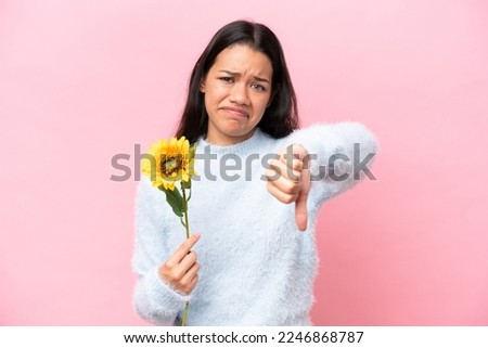 Young Colombian woman holding sunflower isolated on pink background showing thumb down with negative expression