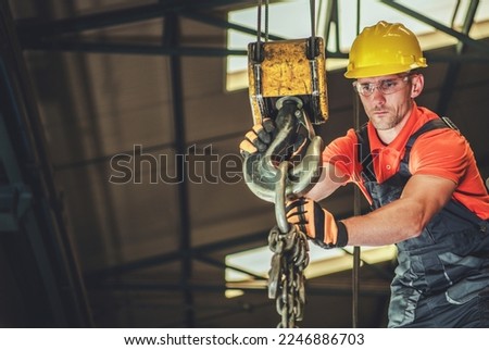 Professional Contractor Lowering the Load Using Large Lifting Hook of Overhead Crane. Indoor Construction Site in the Background. Industrial Theme. Royalty-Free Stock Photo #2246886703