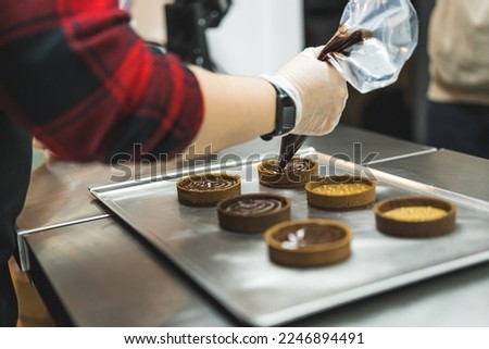 Side view of a baker decorating tarts with chocolate icing from a pastry bag. Blurred foreground. High quality photo