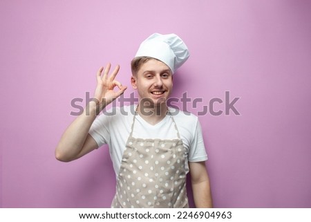 young chef in an apron and a hat shows a gesture of ok with his hand and smiles, a guy baker in uniform on a pink background