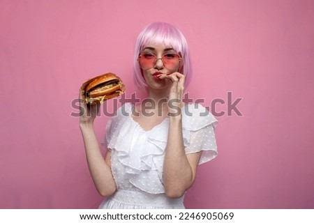 young cute girl with pink short hair holds a big burger in her hands and licks her lips, a woman shows a hamburger on a pink background