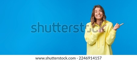 Cheerful hipster girl in yellow hoodie, with blond short haircut, pointing and looking right side with pleased happy smile, promote product or company services, stand blue background.
