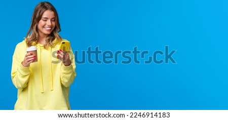 Carefree joyful european female in yellow hoodie, holding take-away coffee and browsing internet in mobile phone, texting friend laughing over funny video or meme, blue background. Royalty-Free Stock Photo #2246914183