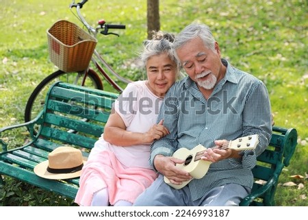 Happy smiling asian senior man and woman sitting on bench playing ukulele and singing a song in garden park outdoor. Musical and relaxation makes lover couple happiness. Health care lifestyle concept.