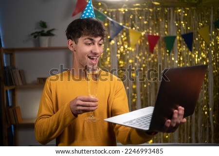 Arabic young man celebrating his birthday through video call virtual party with friends using laptop computer. Holding glass with champagne. Authentic decorated home workplace. Copy space
