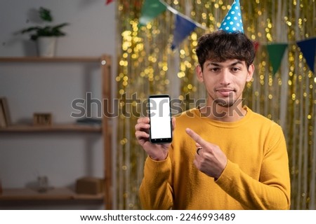 Mockup image of a young man in festive hat pointing finger at a mobile phone with blank white screen, celebrate birthday party at home. Copy space
