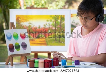 Young Asian boy is looking at example picture on figure stand and using paintbrush to draw various pictures on table outside the room happily, raising teen and adult helps kids school project concept.