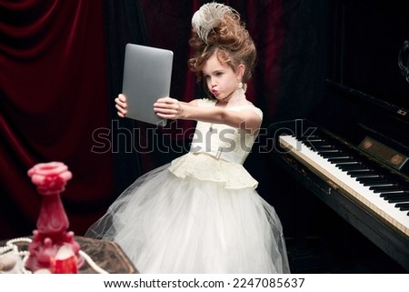 Cute little girl, child in image of medieval royal person in fabulous dress sitting at the piano and taking selfie with tablet. Concept of historical remake, comparison of eras, medieval fashion