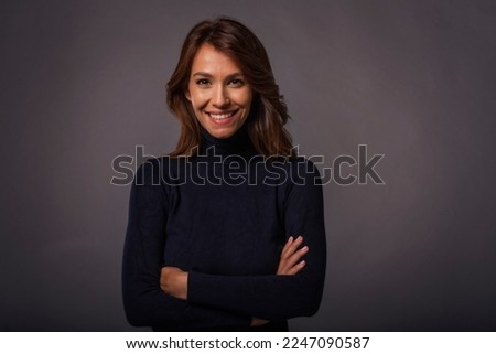 Close-up of an attractive middle aged woman with toothy smile wearing black turtleneck sweater while staning at isolated dark background. Copy space. Studio shot.