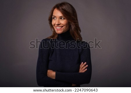 Close-up of an attractive middle aged woman with toothy smile wearing black turtleneck sweater while staning at isolated dark background. Copy space. Studio shot. Royalty-Free Stock Photo #2247090643