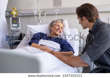 Son visiting sick old mother in hospital. Smiling senior patient in conversation with man. Grandson visiting and cheering his happy granny lying in bed at modern hospital ward. Royalty-Free Stock Photo #2247102511