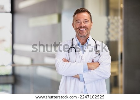 Portrait of happy mature doctor with folded arms standing at hospital hallway. Confident male doctor in a labcoat and stethoscope looking at camera with satisfaction. Smiling general practitioner. Royalty-Free Stock Photo #2247102525