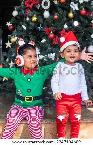 Two Asian Indian siblings in colorful christmas costumes playing around a decorated christmas tree and giving joyful playful expressions