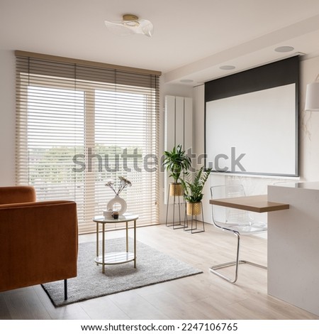Small and elegant living room with projector screen, big window with blinds, cozy velvet sofa, coffee table and plants Royalty-Free Stock Photo #2247106765