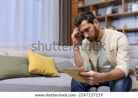 Upset man at home sitting on sofa reading letter with bad news notification in envelope opening, unhappy in living room and sad. Royalty-Free Stock Photo #2247137119