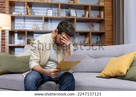 Upset man at home sitting on sofa reading letter with bad news notification in envelope opening, unhappy in living room and sad. Royalty-Free Stock Photo #2247137121