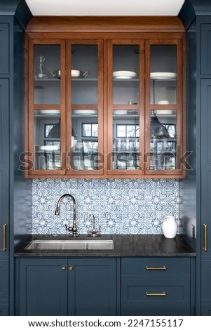 A kitchen sink with a beautiful pattern tiled backsplash with a chrome faucet, black granite countertops, and surrounded by blue and wood cabinets. Royalty-Free Stock Photo #2247155117