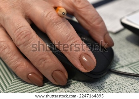 In the close-up picture, a woman's hand, between the fingers of which a pencil is visible, lies on a computer mouse.