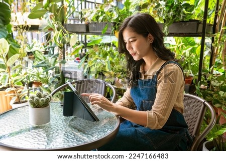 Asian woman owner of a small plant shop sells air purifying plants as a hobby who loves sitting on her tablet and looking at customer orders online at her home plant shop.