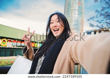 Happy asian young lady taking a selfie photo with an smartphone. Self portrait of chinese smiling woman doing the peace sign after shopping outdoors. High quality photo