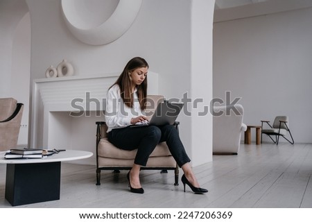 Pensive brunette businesswoman in white shirt and black pants sitting on cozy chair with laptop intense working at office. Serious hispanic female student holds pen, remote learning at home. Student.