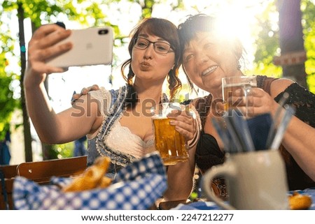 Best friends mother and daughter in Bavarian Tracht making a Selfie with the phone, with mugs of beer in Bavarian beer garden or oktoberfest