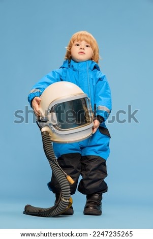 Portrait of little boy, child posing in astronaut costume over blue studio background. Holding giant helmet. Concept of childhood, emotions, lifestyle, fashion, happiness. Copy space for ad