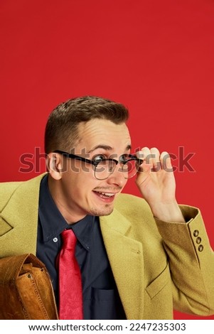Portrait of young man in stylish jacket, glasses and vintage briefcase posing, looking with interest over red background. Job hunting. Emotions, business, occupation, hobby, lifestyle, fashion concept