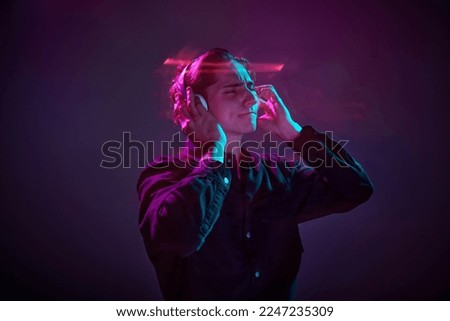 Portrait of young man in casual black shirt posing in headphones isolated over gradient dark purple background in neon mixed light. Concept of human emotions, facial expression, sales, ad, fashion