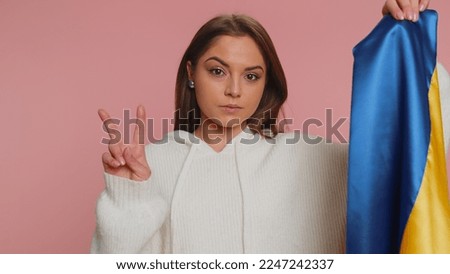 Young pretty woman in casual white sweater holding Ukraine national flag, showing victory sign against war, hoping for success and win. Girl doing peace gesture smiling with kind optimistic expression