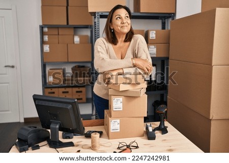 Middle age hispanic woman working at small business ecommerce smiling looking to the side and staring away thinking. 