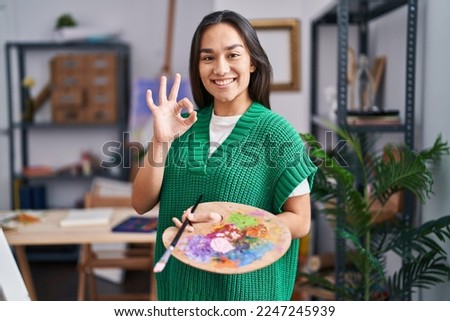 Young south asian woman holding painter palette doing ok sign with fingers, smiling friendly gesturing excellent symbol 