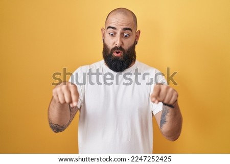 Young hispanic man with beard and tattoos standing over yellow background pointing down with fingers showing advertisement, surprised face and open mouth 