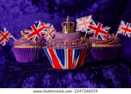 Royal Coronation Cupcakes to celebrate the coronation of King Charles III. Cupcakes decorated with the crown, purple velvet backdrop, union jacks flags, luxury cupcakes on a pedestal.  Royalty-Free Stock Photo #2247268359
