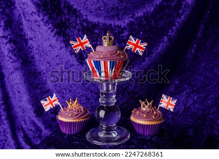 Royal Coronation Cupcakes to celebrate the coronation of King Charles III. Cupcakes decorated with the crown, purple velvet backdrop, union jacks flags, luxury cupcakes on a pedestal.  Royalty-Free Stock Photo #2247268361