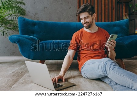 Young man wear red t-shirt hold use work on laptop pc computer mobile phone sit on blue sofa couch stay at home hotel flat rest relax spend free spare time in living room indoors People lounge concept Royalty-Free Stock Photo #2247276167