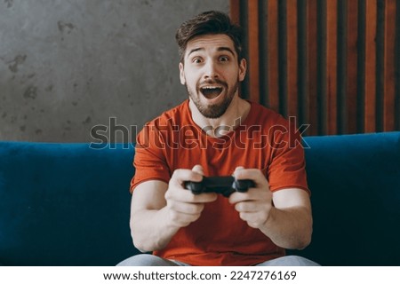 Young excited man wear red t-shirt hold play pc game with joystick console sit on blue sofa couch stay at home hotel flat rest relax spend free spare time in living room indoors People lounge concept