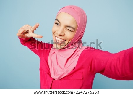 Close up young arabian muslim woman wear pink abaya hijab do selfie shot pov on mobile cell phone blink eye wink isolated on plain light blue cyan background studio. People uae islam religious concept