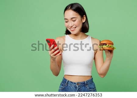 Young cheerful woman wear white clothes hold eat burger use mobile cell phone application isolated on plain pastel light green background. Proper nutrition healthy fast food unhealthy choice concept