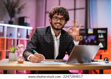 Portrait of attractive confident bearded IT specialist, office manager, wearing suit using laptop making financial report while writing on paper, looking at camera working at night showing sign of ok