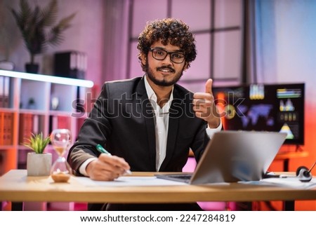 Portrait of attractive confident bearded IT specialist, office manager, wearing suit using laptop making financial report while writing on paper, looking at camera working at night showing thumb up.