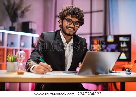 Portrait of attractive confident bearded IT specialist, office manager, wearing suit using laptop making financial report while writing on paper, looking at camera working at night.